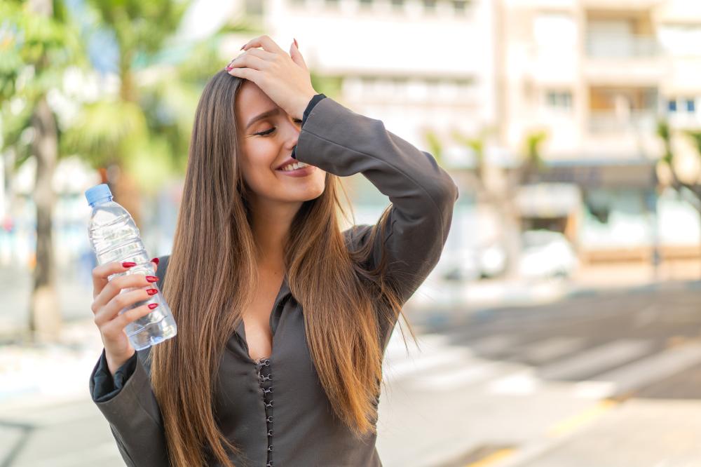 Молодая женщина прикладывает руку ко лбу и держит бутылку с питьевой водой